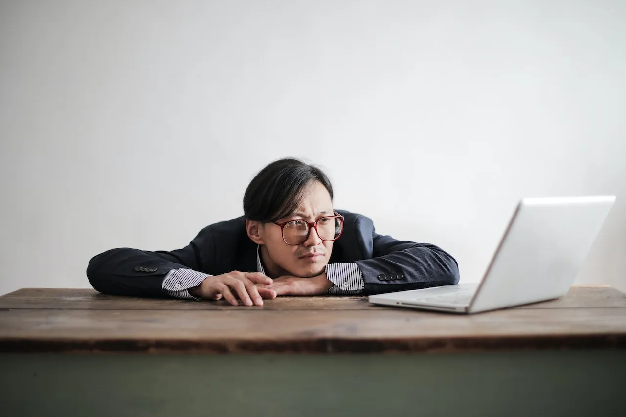 A business man slumped on his desk looking at his screen with a look of boredom on their face